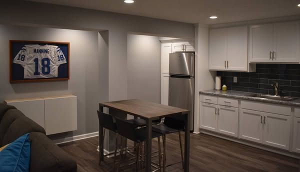 Kitchenette with white cabinets, stainless steel fridge, and small table with two bar stools.