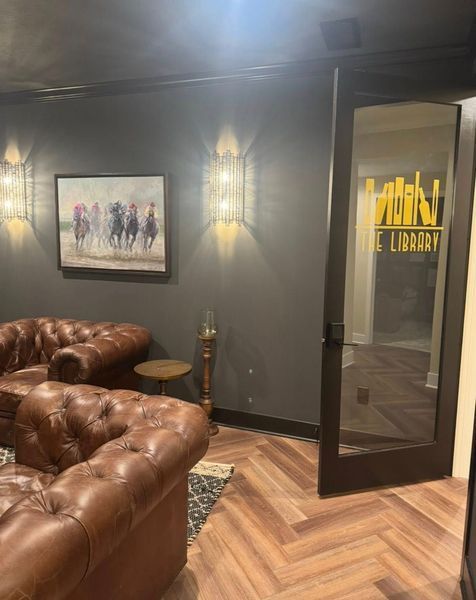 A dark-walled library with leather couches, artwork, and a glass door with 
