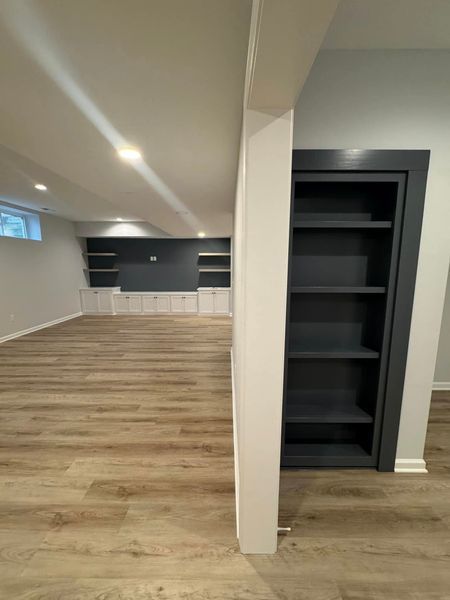 Basement interior with built-in bookshelves painted dark gray. Light wood flooring and white walls.