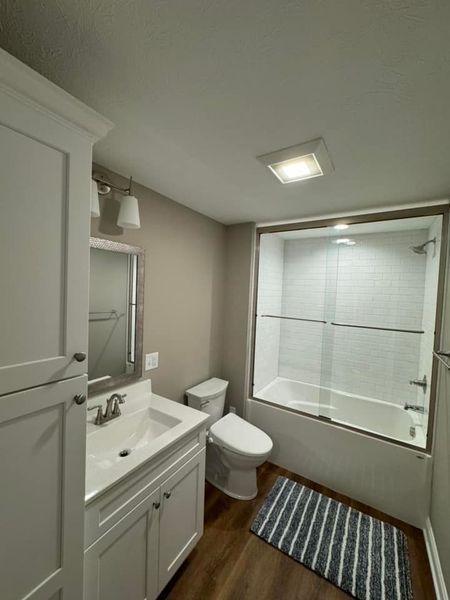 Bathroom with white cabinets, vanity, and toilet. Glass shower doors. Striped rug on wood-look floor.