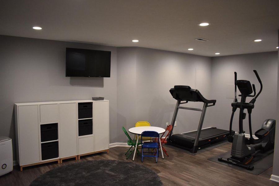 Gym in a gray-walled basement with workout equipment, TV, white cabinet, small table, and chairs.