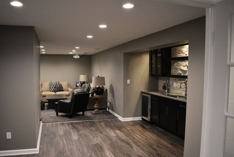 Interior of a basement entertainment area with a bar and seating area; gray walls, wood-look flooring, recessed lighting.