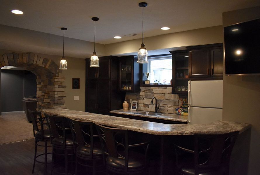 Basement bar with stone archway, dark cabinetry, and pendant lights. A countertop extends with bar stools.