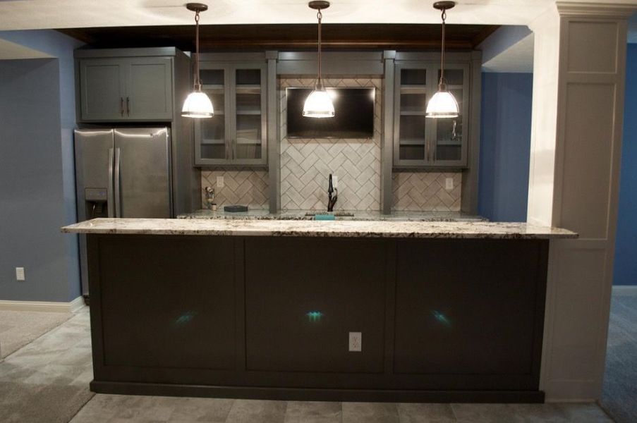 Dark gray basement bar with granite countertop, cabinets, fridge, TV, and pendant lights.