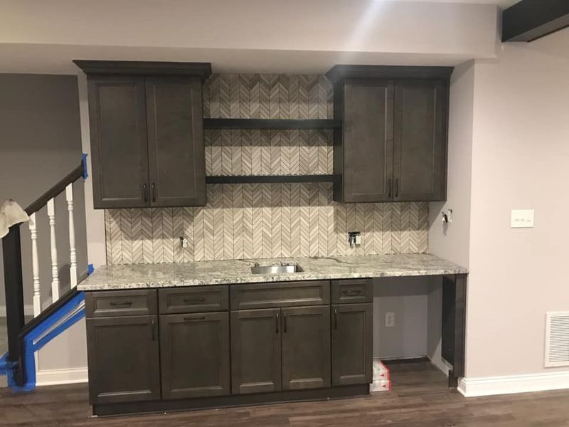 Dark gray cabinetry in a basement bar area with granite countertop, tiled backsplash, and built-in sink.
