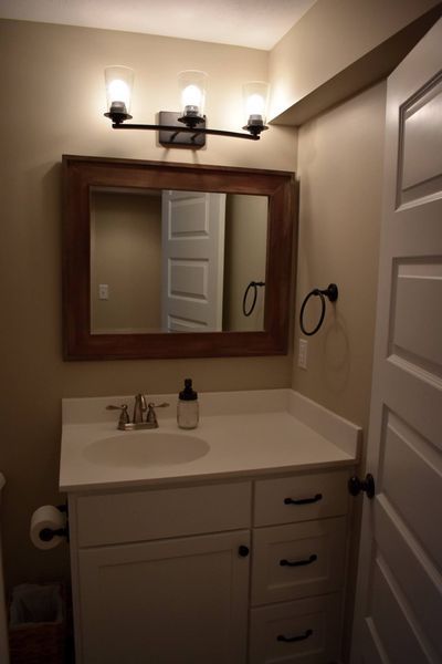 Bathroom with white vanity, mirror in wooden frame, and three-bulb light fixture. Beige walls, white door.