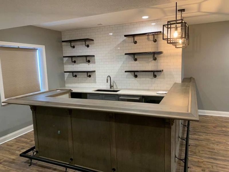 Basement bar with shelves, sink, and pendant light fixture. Wooden bar front, white brick backsplash.
