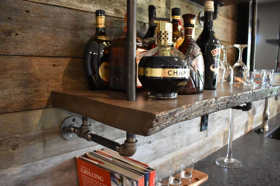 A bar shelf with liquor bottles and glassware, supported by metal pipes, set against wood paneling.