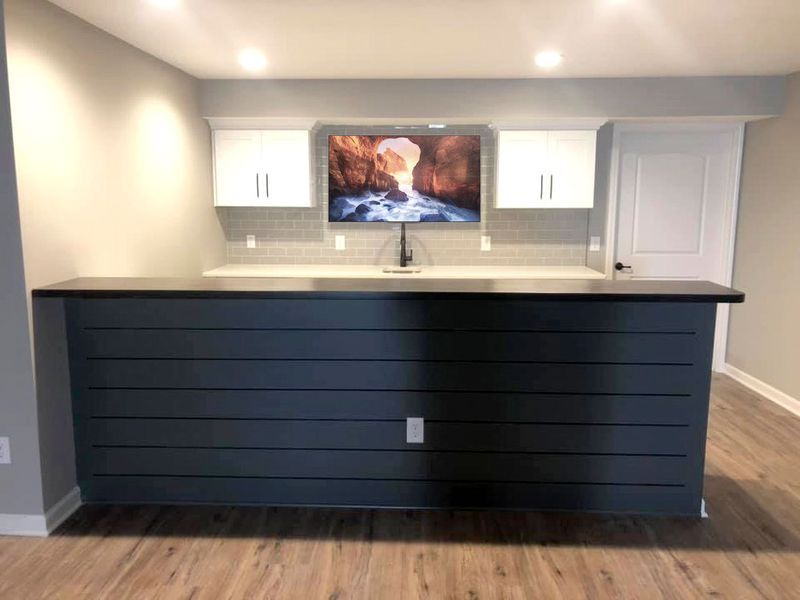 Dark blue shiplap bar with white cabinets, TV, and faucet against a gray wall.