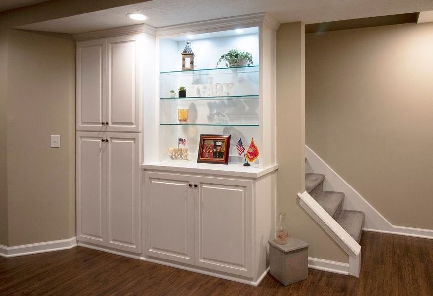Built-in white cabinets with display shelf and staircase in a finished basement. Beige walls, dark wood floor.