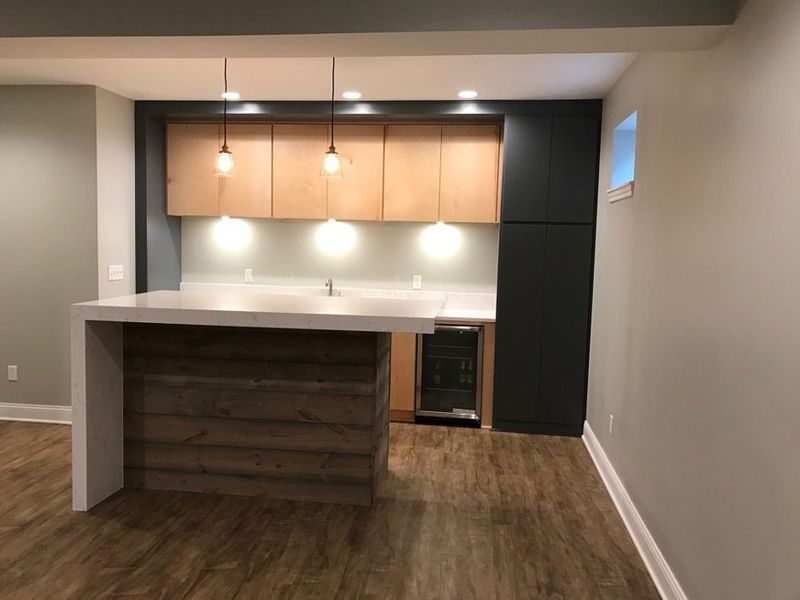 Basement bar with light wood cabinets, gray countertops, and wood-look front; pendant lights.