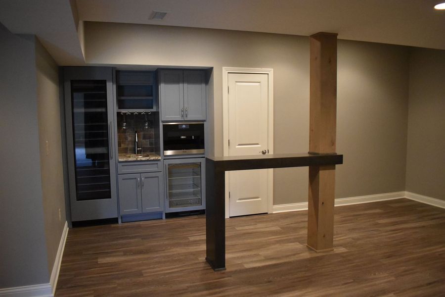 Basement bar area with gray cabinets, a wine fridge, and a dark counter. Wooden floors and post.