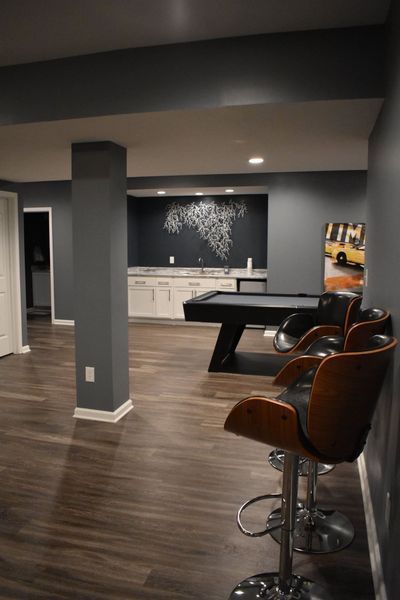 Basement entertainment area with a pool table, bar, and seating. Dark gray walls, wood floors, and neutral accents.