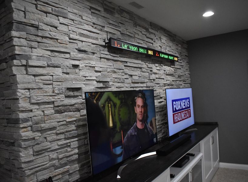 A stone wall with two TVs on a black countertop; one shows a movie scene, the other, a news broadcast. A ticker is above.