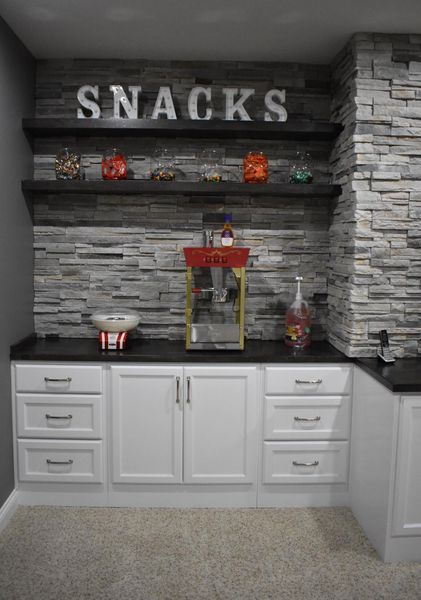 Snack bar with white cabinets, dark countertops, stone accent wall, shelves with snacks, and a popcorn machine.