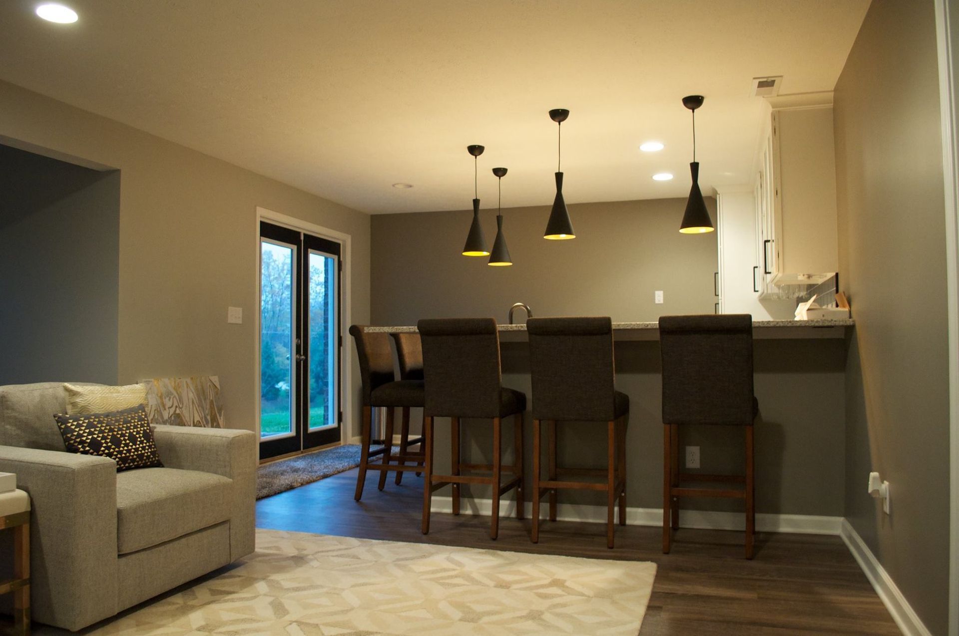 A modern kitchen with a bar, stools, and pendant lights. A seating area with a gray couch is visible on the left.