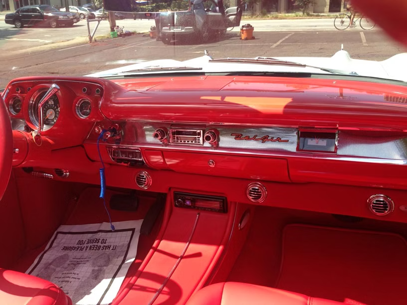 Interior of a vintage red car, showing the dashboard, steering column, and floor mat in a parking lot.