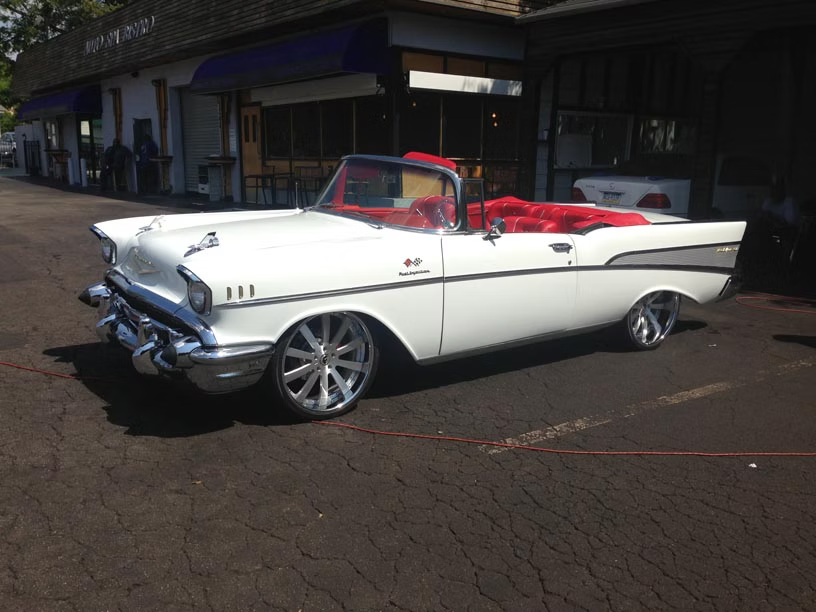 A white 1957 Chevrolet Bel Air convertible with a red interior parked on an asphalt lot.