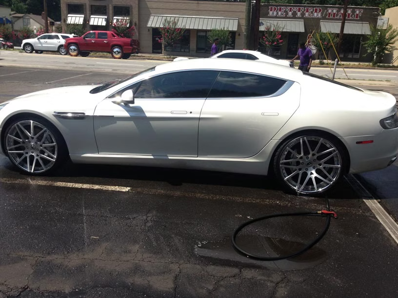 A white Aston Martin Rapide parked on a wet asphalt surface in front of a small shopping center.