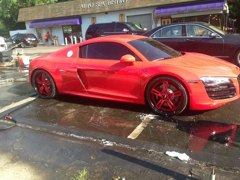 A bright red Audi R8 parked in front of a building with a purple awning, with wet pavement and another car visible nearby.