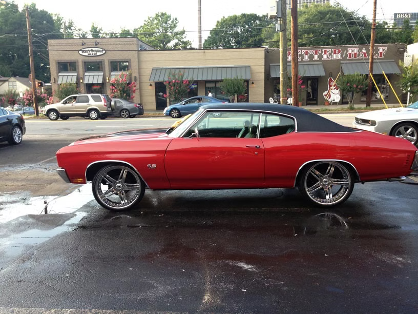 A red vintage Chevrolet Chevelle with a black vinyl roof and large chrome rims parked on a wet asphalt lot.