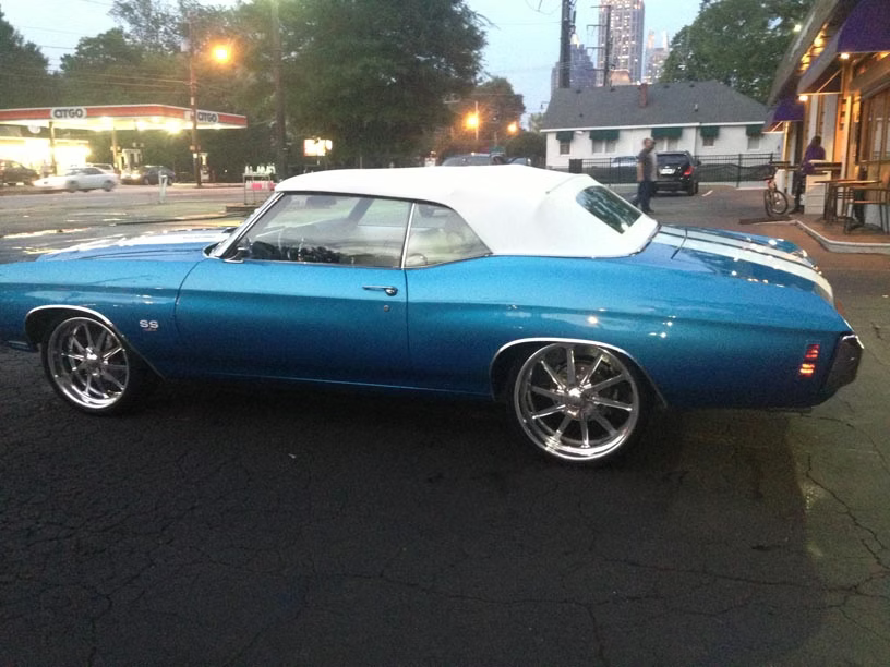 A bright blue Chevrolet Chevelle convertible with a white top and custom wheels, parked on an asphalt street at dusk.