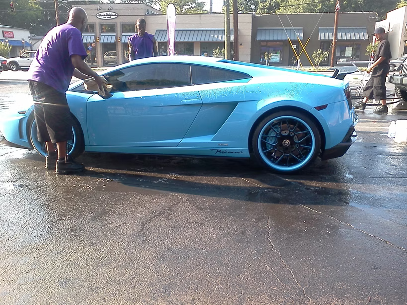 A person washes a bright blue Lamborghini sports car at an outdoor car wash station.