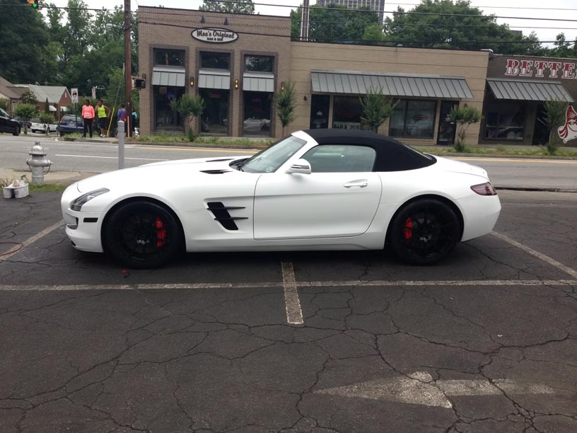 A white Mercedes-Benz SLS AMG convertible with black wheels parked in an asphalt lot in front of commercial buildings.