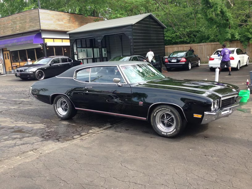 A black vintage Buick muscle car parked in a paved lot in front of a small building, with other cars in the background.