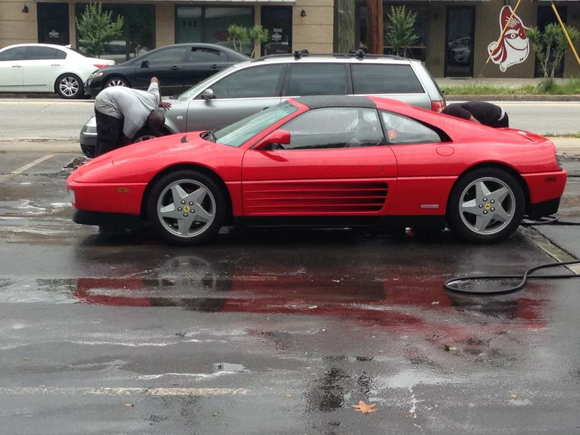 A red Ferrari sports car parked on a wet parking lot being cleaned by two individuals.