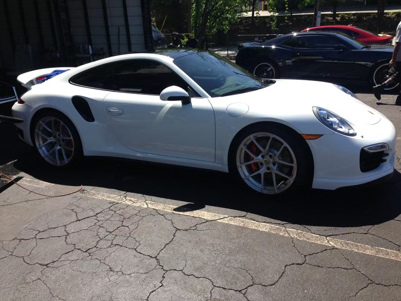A white Porsche 911 Turbo parked on a cracked asphalt lot on a sunny day.