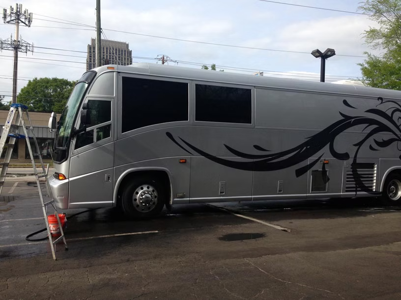 A silver tour bus with black floral graphics parked in a lot, with a tall ladder resting against the front windshield.