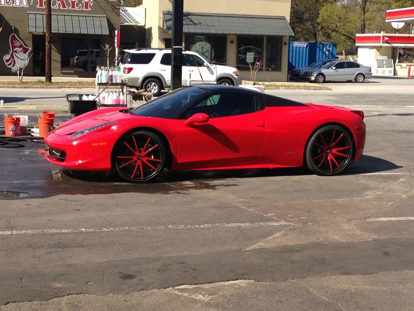 A red Ferrari sports car with custom red-rimmed wheels parked in a parking lot on a sunny day.