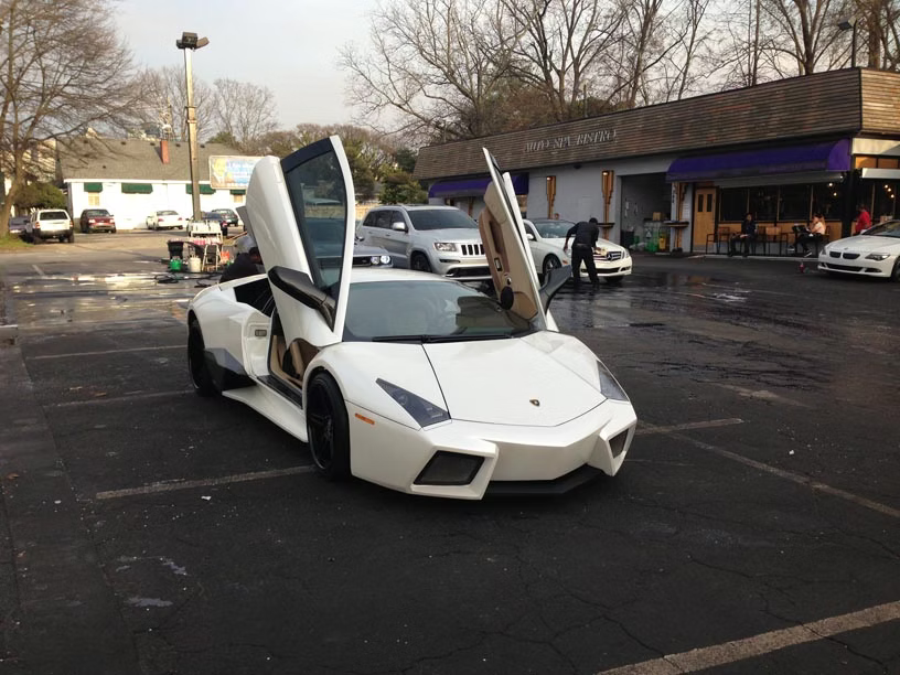A white Lamborghini with its scissor doors open is parked in an outdoor lot during the day.