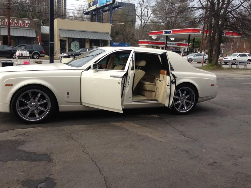 A white Rolls-Royce with its rear-hinged doors open, parked in a parking lot near a gas station.