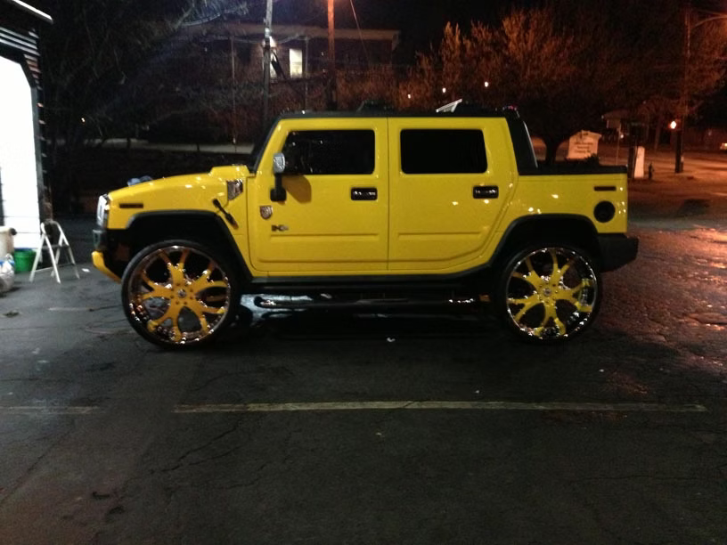 A yellow Hummer H2 SUT with large, gold-accented custom wheels parked in a parking lot at night.
