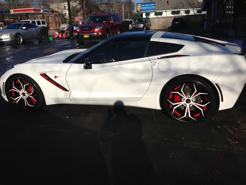A white Chevrolet Corvette with custom red and white spoke rims parked on asphalt at a car wash.