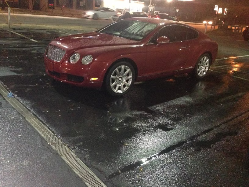 A red Bentley Continental GT coupe parked in a wet asphalt parking lot at night.