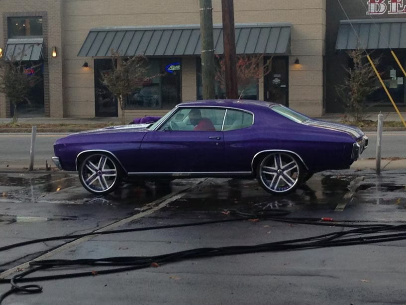 A purple vintage muscle car with large, custom chrome wheels parked on a wet asphalt lot in front of a building.