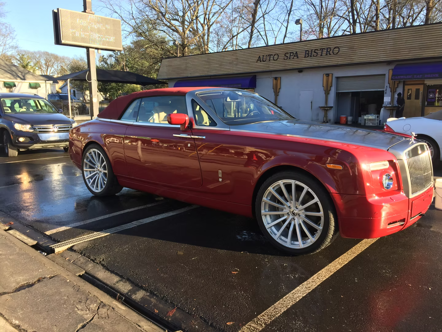 A red Rolls-Royce convertible parked in a lot in front of a building on a sunny day.