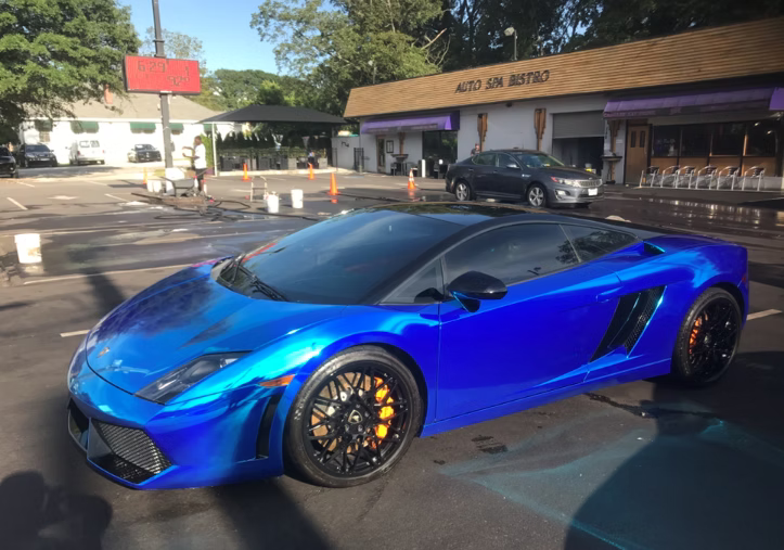 A metallic blue Lamborghini Gallardo parked in front of an auto shop on a sunny day.