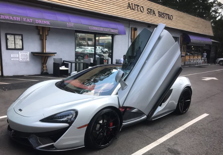 A silver McLaren sports car with butterfly doors open, parked in front of the Auto Spa Bistro.