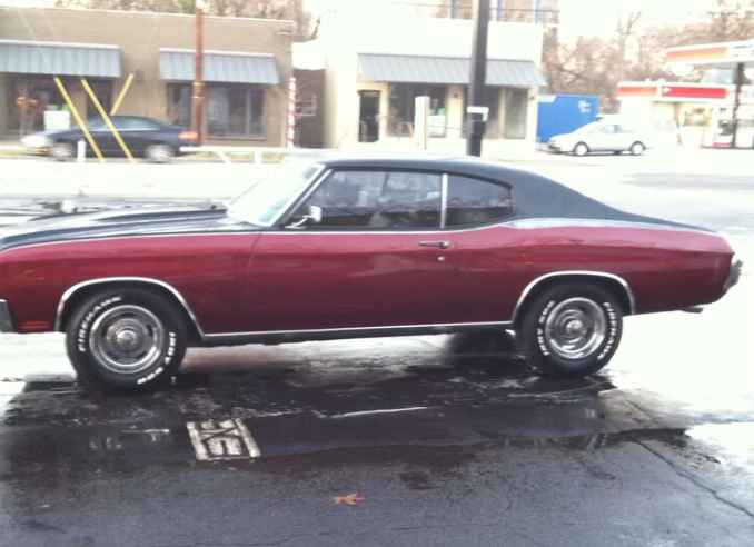 A deep red classic Chevrolet Chevelle with a black vinyl top and racing stripes parked on a wet street.