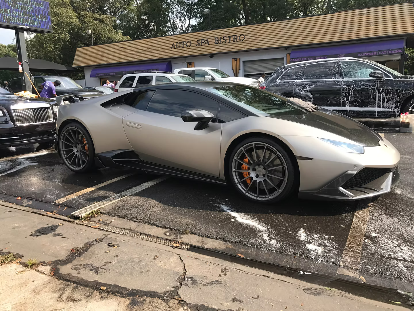 A metallic champagne-colored Lamborghini Huracán parked at an outdoor car wash, with soap suds on the asphalt.