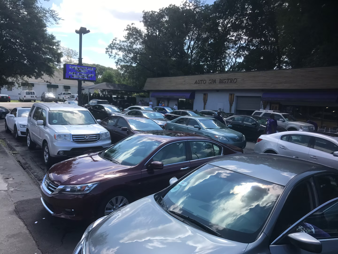 A crowded parking lot filled with various sedans and SUVs in front of a commercial building with a digital sign.