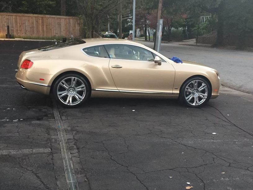 A gold Bentley coupe parked on a paved lot beside a road.