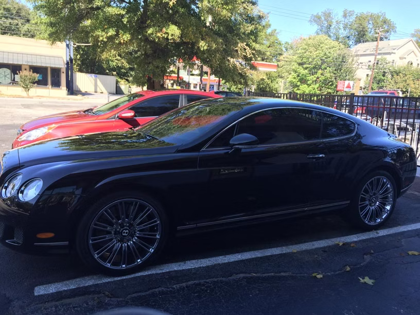 A sleek black Bentley coupe parked outdoors next to a red sedan on a sunny day.