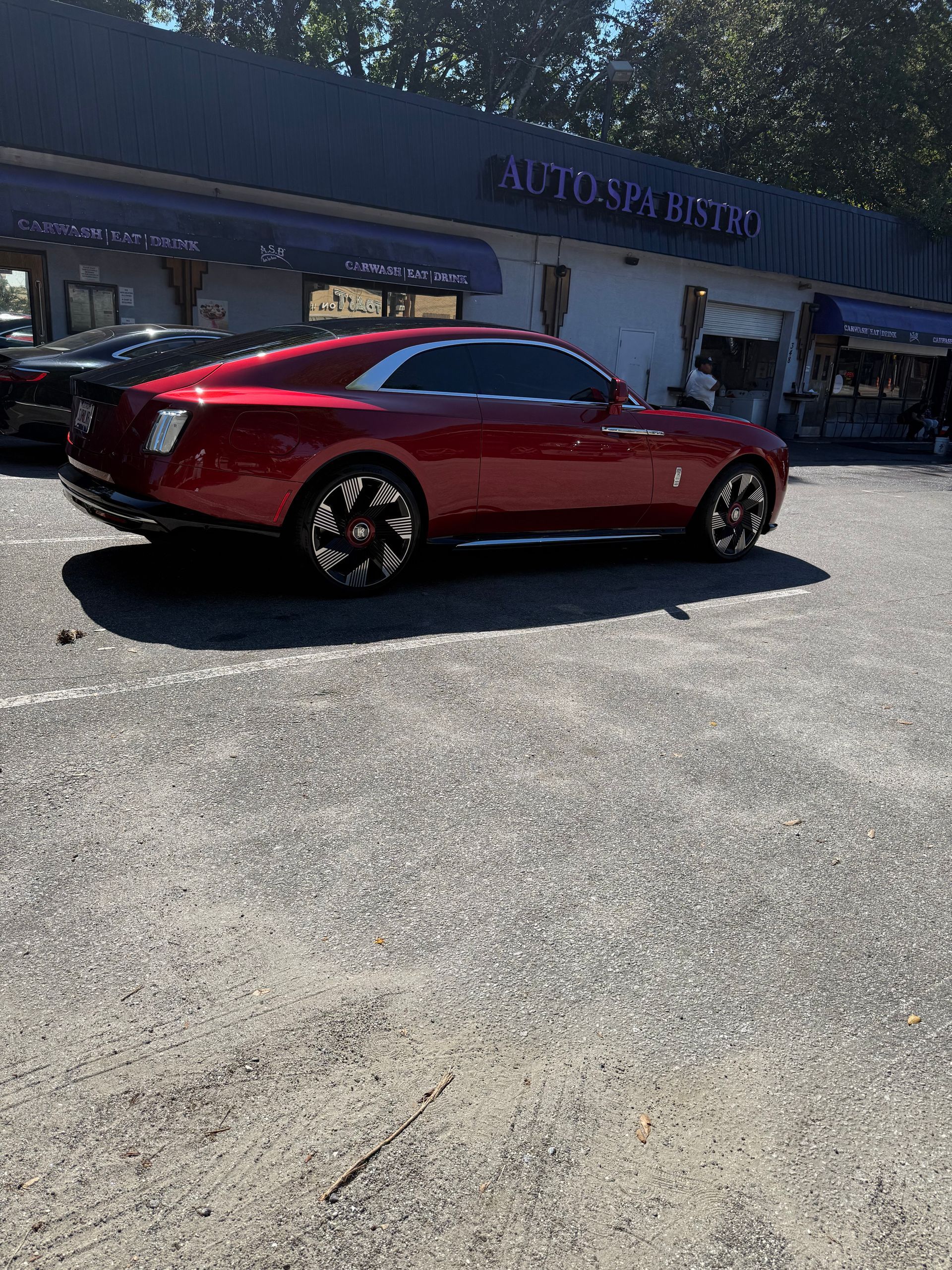 A cherry-red Rolls-Royce Spectre parked on a gravel lot in front of an Auto Spa building.
