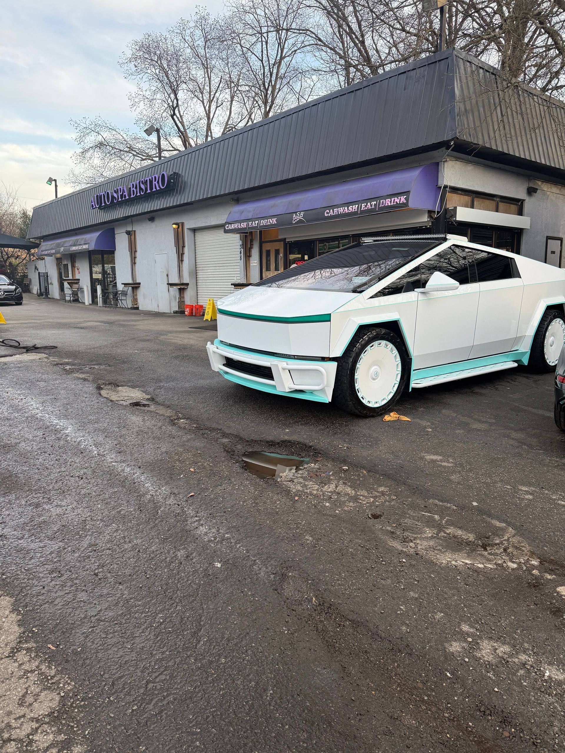 A white Tesla Cybertruck with a custom teal lower trim parked in front of a commercial building on a cloudy day.