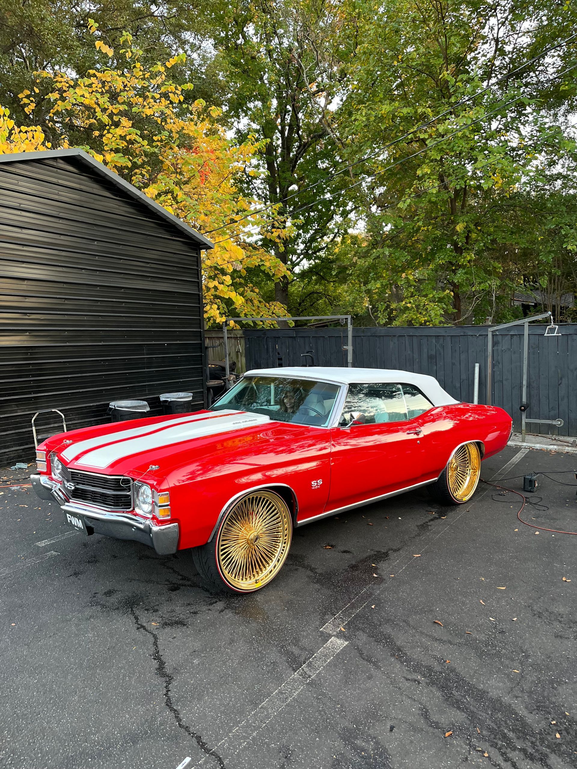 A bright red convertible classic car with white racing stripes and large, gold-rimmed wheels parked on an asphalt lot.
